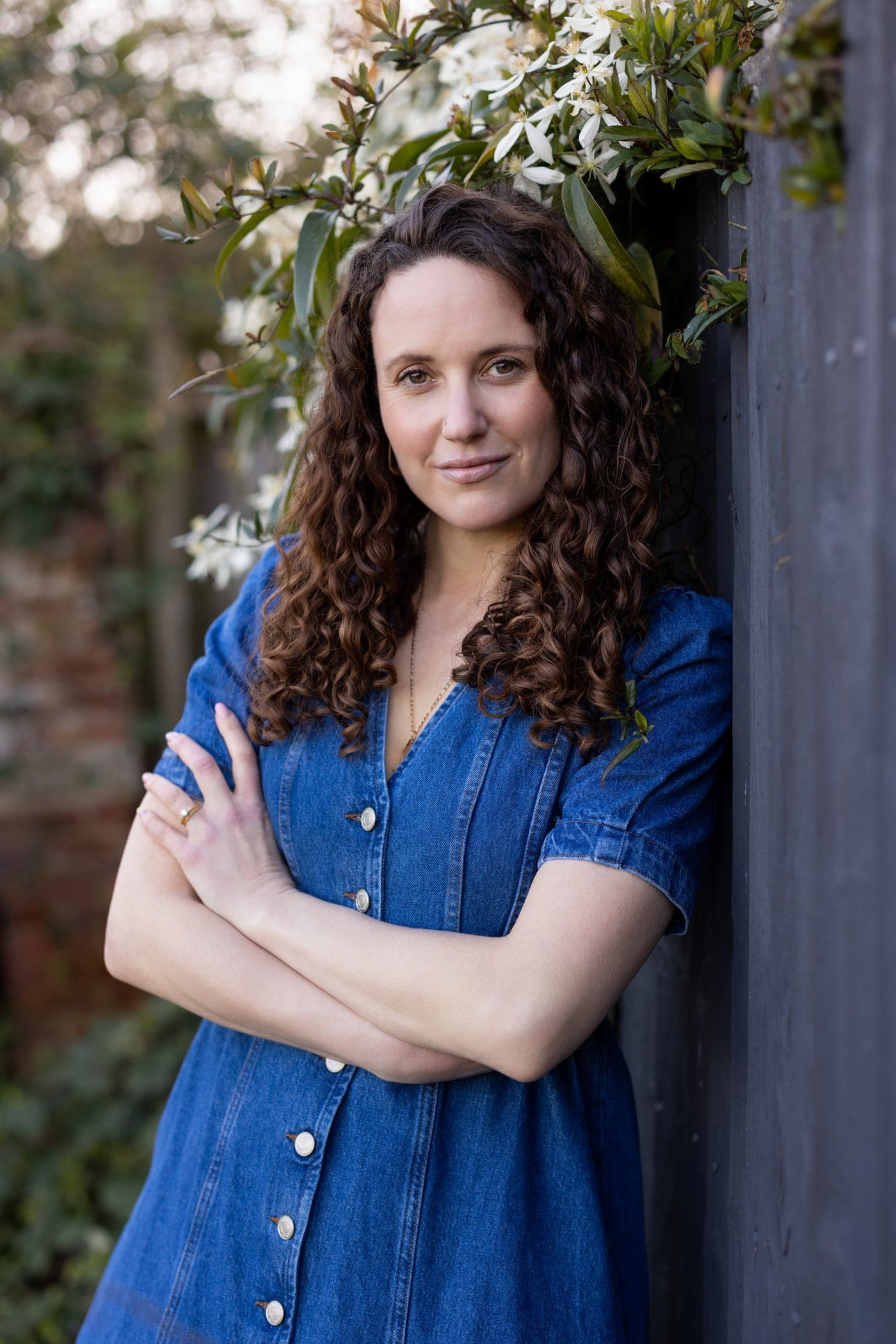 Mrs Gill standing in a garden beside white blossom, wearing a blue denim dress.